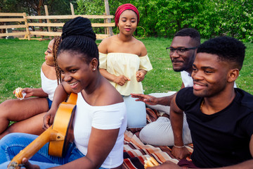 Group of smiling happy afro-american friends friends playing guitar outdoors picnic in the campaign