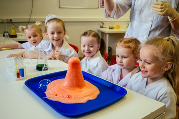group of school girl kids with teacher in school laboratory making experiment observing the chemical reaction with the dye with vinegar and soda volcano wearing white gown uniform glass © yurakrasil