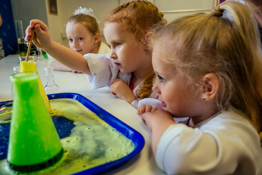 Group Of School Girl Kids With Teacher In School Laboratory Making Experiment Observing The Chemical Reaction With The Dye With Vinegar And Soda Volcano Wearing White Gown Uniform Glass