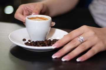 Girl hand holding espresso coffee cup on the desk in a bar. On saucer is blurred roasted coffee beans. Enjoying coffee concept. Close up, selective focus