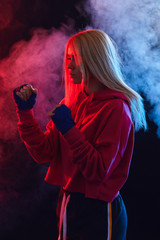 Female blonde boxer in pink hoodie prepares to punch at a boxing studio. Woman boxer in motion over black smoky background