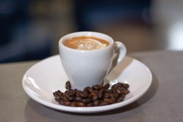 Espresso coffee in a white cup and roasted coffee beans on a saucer on wooden desk in a bar. Brown coffee with milk and bubble. Close up, selective focus