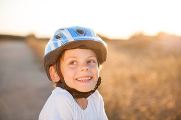 portrait of happy smiling cute little caucasian kid in blue sport safe helmet and white shirt on summer sunset outdoors