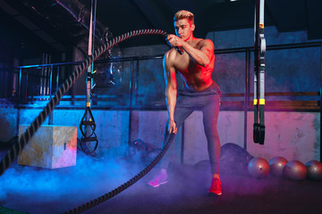 Handsome muscular bare-chested gym man with battle ropes exercise in the fitness gym. Cross fit training workout.