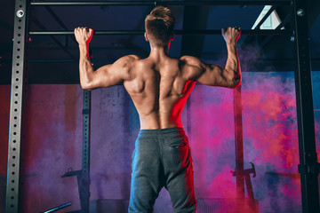 Young bodybuilder training in the gym exercising Wide-Grip Pull-Up in dark gym with blue lightning,...