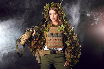 Heavily armed female soldier in battle helmet and ghillie suit holding assault rifle isolated on dark smoky battlefield. Paint ball and laser tag sport games
