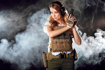 Fototapeta na wymiar Armed beautiful blonde woman wearing protective headphones and plate carrier, shoots with gun at a target in the darkness with smoke clouds