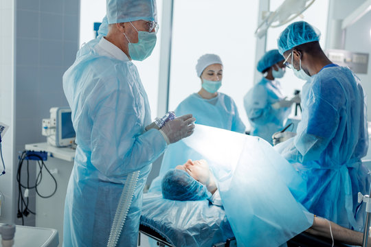 Male Patient Lying On An Operating Table With Surgeons In A Surgery Room. Medicine Concept