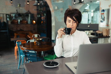 A middle-aged woman sits in a cafe, drinks coffee and works at a computer
