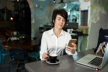 A middle aged woman sits in a cafe and works at a laptop.