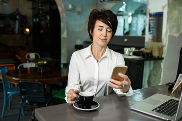 A middle-aged woman sits in a cafe, drinks coffee and works at a computer. Woman holding a phone