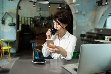 A middle-aged woman sits in a cafe, drinks coffee and works at a computer. Woman holding a phone