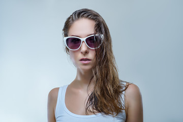 Naklejka premium Close up portrait of beautiful young woman in fashionable white sunglasses and white shirt is posing stylishly isolated on gray background in a studio