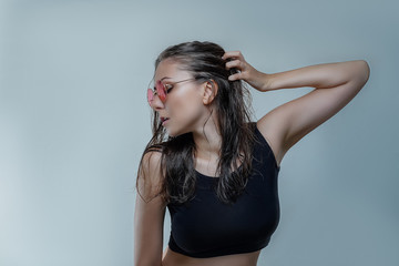Fototapeta premium Portrait of beautiful young woman in fashionable round pink sunglasses and black shirt with wet hair is posing stylishly in profile isolated on gray background close up
