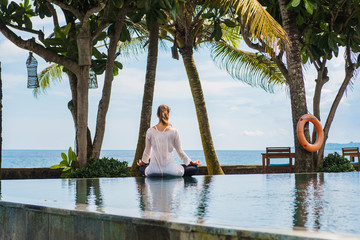 Woman from the back in sport suit is sitting in lotus pose and meditating on the edge of the pool on the coast of ocean in tropical nature with palm trees