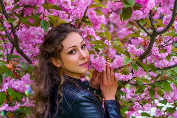 Naklejka premium Portrait of beautiful woman with curly hair in white dress and black leather jacket is smiling in blooming pink flowers. Fashion photo shoot in the garden