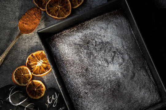 Blacha ciasta czekoladowego, posypanego cukrem pudrem. Dookoła suszone pomarańcze i łyżka kakao.