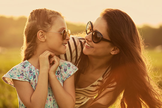 Happy fashion kid girl embracing her mother in trendy sunglasses and looking each other with love on nature background. Closeup portrait