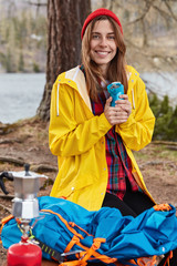 Outdoor shot of European woman with cheerful expression, stands on knees at ground in forest near river, holds flask of hot drink, wears red hat and yellow raincoat, coffee maker on camping stove