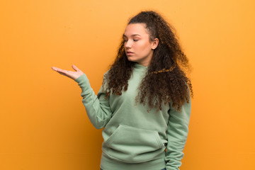 Teenager girl over ocher wall holding copyspace with doubts