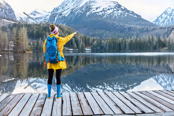 Horizontal shot of female traveler in yellow raincoat and blue rubber boots stands in front of mountain landscape, poses on pier near lake with turquoise water, points into distance, enjoys freedom