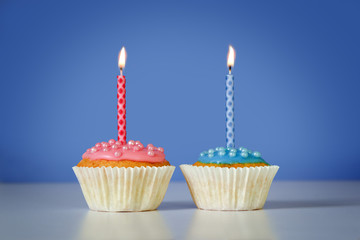 two muffins or cupcakes with pink and blue burning candles against a blue background, birthday concept with copy space