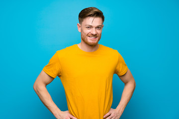 Redhead man over blue wall posing with arms at hip and smiling