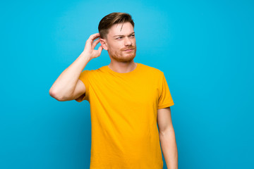 Redhead man over blue wall having doubts while scratching head