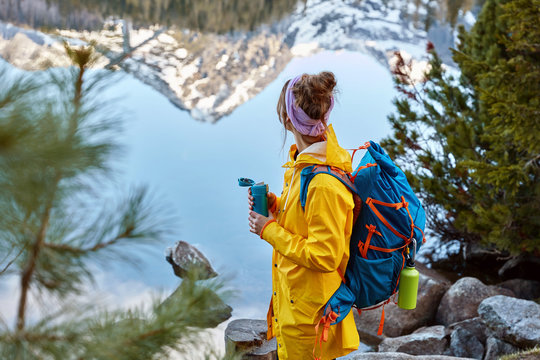 Hiking woman stops near lake in mountains, carries backback, holds thermos of hot beverage, explores something new, wears raincoat stands sideways at camera. Leisure, vacation, adventure concept