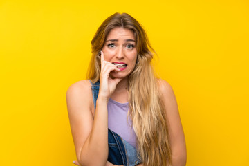 Young blonde woman with overalls over isolated yellow background nervous and scared