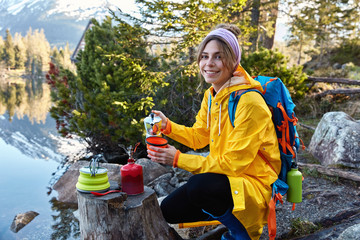 Glad female tourist pours hot aromatic drink from coffee maker, has camping adventure, wears yellow raincoat and headband, wanders in forest near mountain lake, smiles at camera. Tourism concept