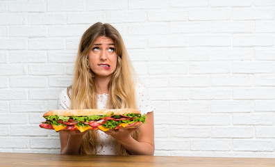 Young blonde woman holding a big sandwich having doubts