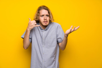 Blonde man over isolated yellow background making phone gesture and doubting