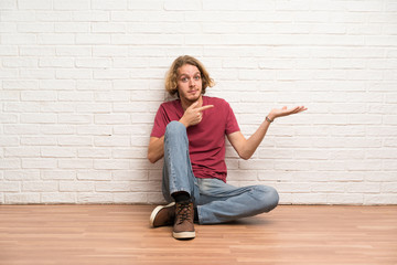 Fototapeta na wymiar Blonde man sitting on the floor holding copyspace imaginary on the palm to insert an ad