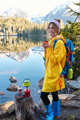 Pleased young woman smiles gently, drinks hot beverage, wears raincoat and rubber boots, enjoys sunny weather after rain, uses tourist equipment, poses near moutain lake, has spare time for rest © WHstudio Leushin N