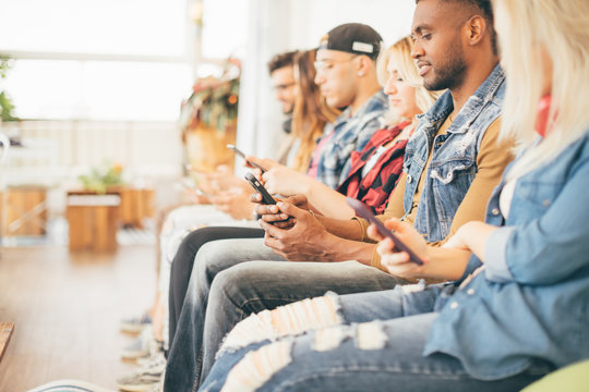 Picture of multiracial group of young people sitting with smartphones in their hands, looking into screens and smiling - technology and social media addiction concept