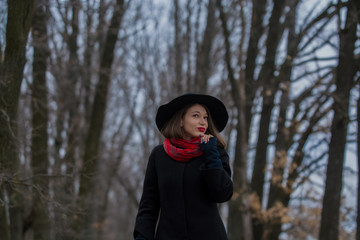 Fototapeta na wymiar The girl in a black coat, a shovel hat and with red lipstick on lips, walks in the park. Style and modern fashion.