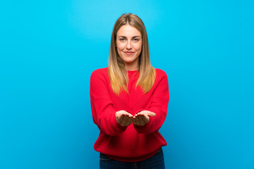Woman with red sweater over blue wall holding copyspace imaginary on the palm to insert an ad