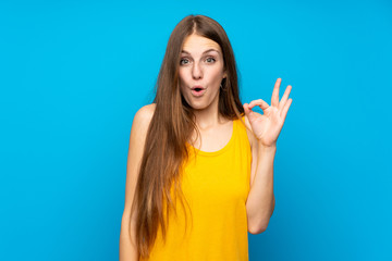 Young woman with long hair over isolated blue wall surprised and showing ok sign