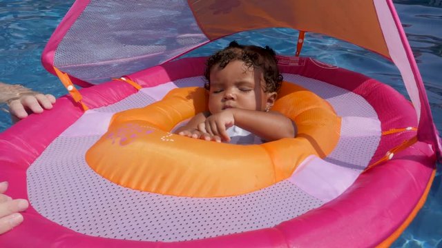 baby girl alseep in floaty in pool. A baby multi ethnic girl is a sleeping in pool floaty as dad moves it around so we can she her face.