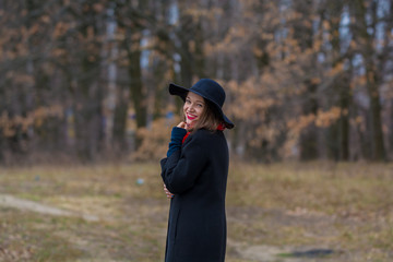 The girl in a black coat, a shovel hat and with red lipstick on lips, walks in the park. Style and modern fashion.