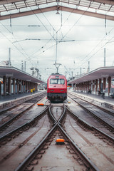 Frontal vertical view of a modern red locomotive cabin arriving into railway station depot; contemporary electric high-speed suburban train leaving the platform with tracks junction in front