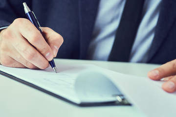 Hand of businessman in suit filling and signing with blue pen partnership agreement form clipped to pad closeup. Management training course, some important document, team leader ambition concept.