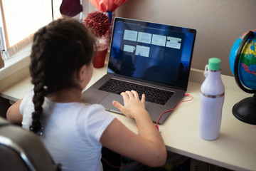 Children girl typing on laptop keyboard on his homework table with thermo bottle and other supplies