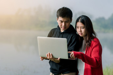 Lovely Asian couple is bright in the tourist area. Laptop computer hands in the hands of the road amid the fog The back is a lake And the sun As happy as looking at the computer screen smiling happily