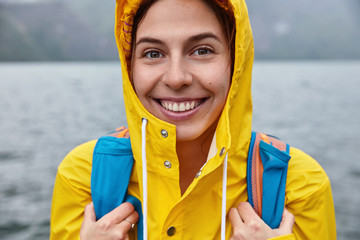 Cropped image of happy woman wears yellow anorak, hood on head, smiles happily, carries luggage, spends holidays in paradise place, majestic scenery water view in background. Tour and people © WHstudio Leushin N