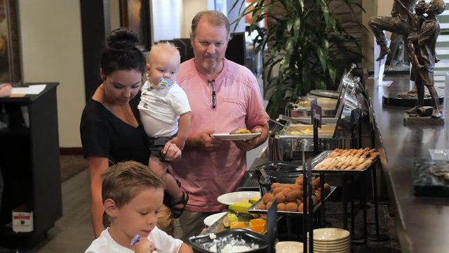 young mom dishes kids food from breakfast buffet line. A man waits in line for a hotel breakfast buffet as mother with kids dish up food.