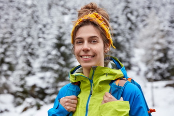 Happy tourist poses on snowy mountain top, enjoys winter day trekking, wears yellow headband, casual jacket, carries backpack, enjoys outdoor activities and active lifestyle, hikes on snow trail