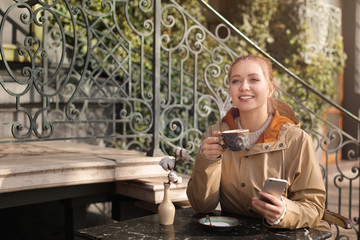 Young woman enjoying tasty coffee while using mobile phone at table outdoors