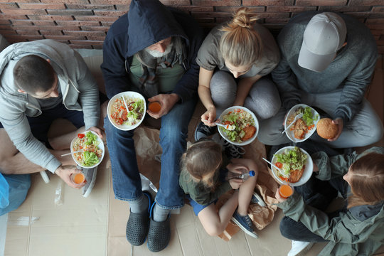 Poor people with plates of food sitting at wall indoors, view from above